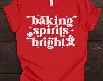 May include: A red t-shirt with the phrase "baking spirits bright" in white, featuring a cookie and gingerbread man graphic. The shirt is a festive holiday design.