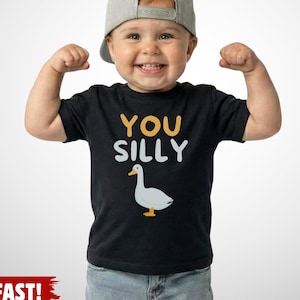 K&ouml;nnte beinhalten: Ein schwarzes kurz&auml;rmeliges T-Shirt mit dem Text YOU SILLY in gelben und wei&szlig;en Buchstaben &uuml;ber einer Grafik einer wei&szlig;en Gans mit orangefarbenem Schnabel und F&uuml;&szlig;en. Das T-Shirt besteht aus Baumwolle und ist f&uuml;r Kleinkinder konzipiert.