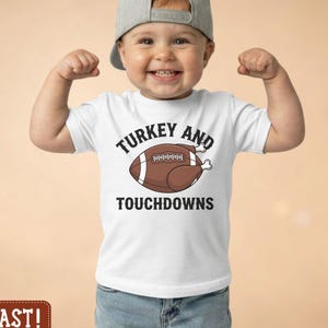 May include: A white short-sleeved t-shirt featuring a graphic of a brown football shaped like a roasted turkey with the text Turkey and Touchdowns printed in black. The shirt is paired with blue denim jeans and a grey baseball cap.