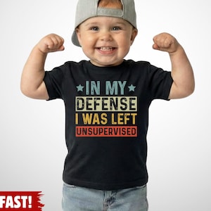 May include: A black t-shirt with the text "IN MY DEFENSE I WAS LEFT UNSUPERVISED" in a colorful, vintage-style font. The shirt is worn by a smiling child wearing a gray cap and blue jeans.