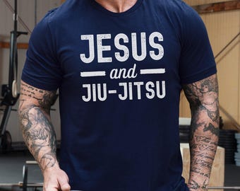 May include: A navy blue short-sleeved t-shirt featuring the white distressed-style text "JESUS and JIU-JITSU" printed across the chest. The shirt has a crew neckline and a standard fit, showcasing a clean design with bold, legible lettering.