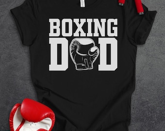 May include: A black short-sleeved t-shirt featuring the white text BOXING DAD with a graphic of a boxing glove replacing the letter A. The shirt is displayed with red boxing gloves and a jump rope with red handles placed around the garment.