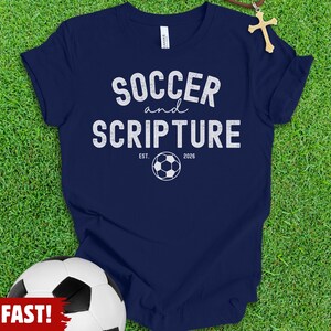 May include: A navy blue short-sleeve t-shirt featuring the white text SOCCER and SCRIPTURE with a soccer ball graphic and the text EST. 2026. A small wooden cross pendant on a brown cord is positioned near the neckline of the shirt.