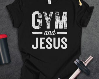 May include: A black short-sleeved t-shirt featuring the white distressed text GYM and JESUS centered on the chest. The shirt is displayed alongside a black shaker bottle, a metal dumbbell, and a gold-colored cross pendant on a brown cord.