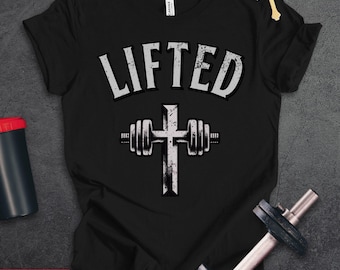 May include: A black short-sleeve t-shirt featuring the word LIFTED in a distressed white font above a graphic of a cross combined with a barbell. The design is centered on the chest. The shirt is displayed alongside a black shaker bottle and a dumbbell.