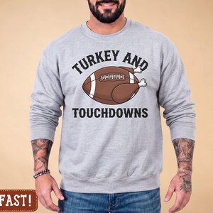 Op de afbeelding: Een asgrijs sweatshirt met ronde hals en een afbeelding van een bruine American football in de vorm van een gebraden kalkoen met witte botten aan de uiteinden. De tekst boven en onder de afbeelding luidt "TURKEY AND TOUCHDOWNS" in zwarte letters.