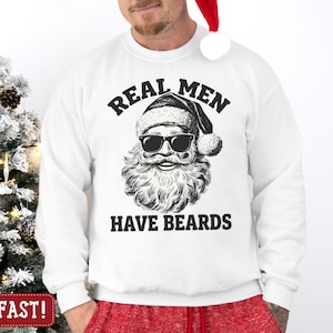 May include: A white long-sleeve sweatshirt featuring a black graphic print of Santa Claus wearing sunglasses and a Santa hat. The text above the graphic reads REAL MEN and the text below the graphic reads HAVE BEARDS in a bold, black font.