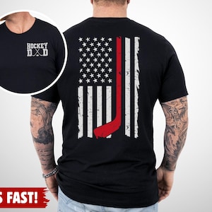 May include: A black t-shirt featuring a white and red graphic on the back of a distressed American flag with a red hockey stick integrated into the design. The front chest area displays the white text HOCKEY DAD with two crossed hockey sticks.