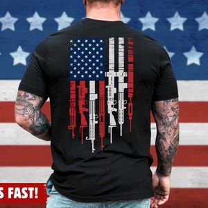 May include: Black t-shirt featuring a stylized American flag design with rifles replacing the stripes. The flag's stars are in the upper left corner. The shirt has the text "BLACK" in a circle on the upper right. The shirt is made of a soft material.