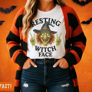 May include: White t-shirt with a witch graphic and the text "RESTING WITCH FACE". The person is wearing a black and orange striped cardigan and blue jeans. Halloween-themed apparel.