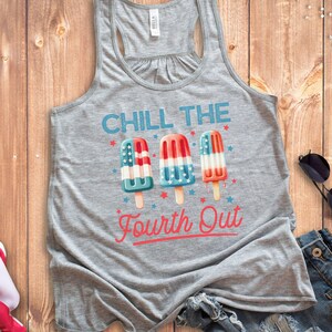 May include: Athletic heather tank top with the text "CHILL THE Fourth Out" and three popsicle graphics in red, white, and blue. The tank top is on a wooden surface with a folded American flag and sunglasses.