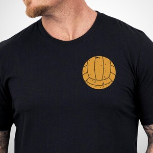 Puede incluir: Una camiseta negra de manga corta con un gr&aacute;fico de un bal&oacute;n de waterpolo de color amarillo mostaza de estilo vintage en el pecho izquierdo. El dise&ntilde;o del bal&oacute;n tiene un aspecto desgastado y texturizado con l&iacute;neas y motas marr&oacute;n oscuro.