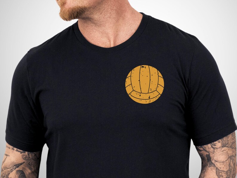 Puede incluir: Una camiseta negra de manga corta con un gr&aacute;fico de un bal&oacute;n de waterpolo de color amarillo mostaza de estilo vintage en el pecho izquierdo. El dise&ntilde;o del bal&oacute;n tiene un aspecto desgastado y texturizado con l&iacute;neas y motas marr&oacute;n oscuro.