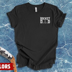 May include: A dark grey heather t-shirt featuring a white graphic on the left chest. The graphic displays the text HOCKEY DAD in a block font, with two crossed hockey sticks positioned between the letters D and A, replacing the letter A.