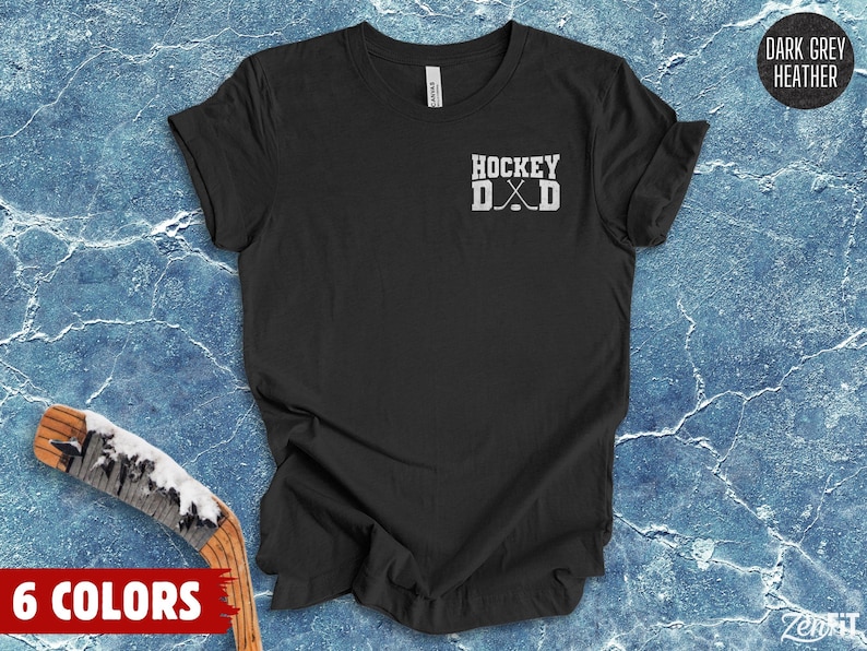 May include: A dark grey heather t-shirt featuring a white graphic on the left chest. The graphic displays the text HOCKEY DAD in a block font, with two crossed hockey sticks positioned between the letters D and A, replacing the letter A.