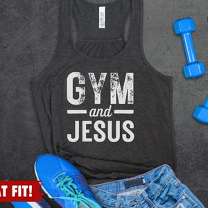 May include: A dark grey heather racerback tank top with the white distressed text GYM and JESUS printed on the front. The garment is displayed alongside two blue dumbbells, a blue athletic shoe with teal laces, and blue denim shorts.