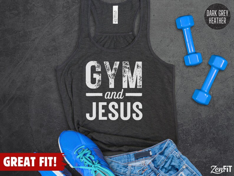 May include: A dark grey heather racerback tank top with the white distressed text GYM and JESUS printed on the front. The garment is displayed alongside two blue dumbbells, a blue athletic shoe with teal laces, and blue denim shorts.