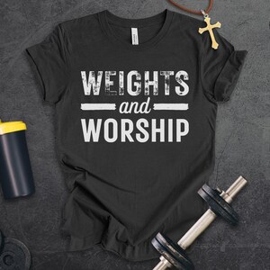 May include: A dark grey heather t-shirt featuring the white text "WEIGHTS and WORSHIP" in a distressed font. The shirt is displayed alongside a black shaker bottle with a yellow lid, a metal dumbbell, and a gold cross pendant on a brown cord.