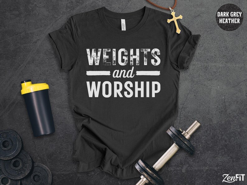 May include: A dark grey heather t-shirt featuring the white text "WEIGHTS and WORSHIP" in a distressed font. The shirt is displayed alongside a black shaker bottle with a yellow lid, a metal dumbbell, and a gold cross pendant on a brown cord.