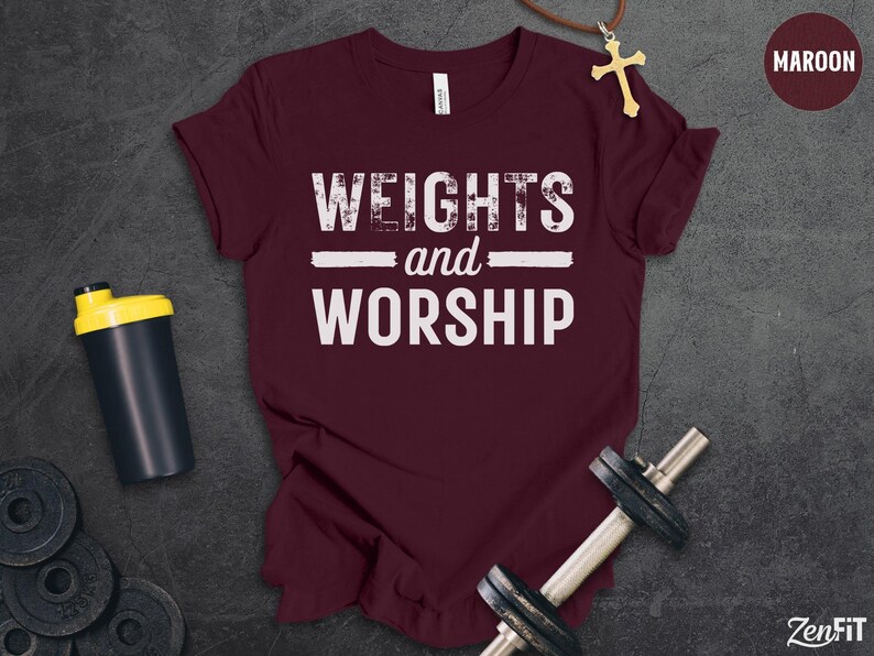 May include: A maroon short-sleeved t-shirt featuring the white text "WEIGHTS and WORSHIP" printed on the front. The shirt is displayed alongside a black shaker bottle, a metal dumbbell, and a gold cross necklace on a dark textured surface.