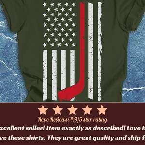 May include: A military green t-shirt featuring a distressed white and red graphic of the American flag. The flag design incorporates a red hockey stick silhouette integrated into the stripes. The shirt has a crew neckline and short sleeves.