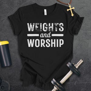 May include: A black t-shirt featuring the white distressed text "WEIGHTS and WORSHIP" centred on the chest. The shirt is accompanied by a black and yellow shaker bottle, a metal dumbbell, and a gold cross pendant on a brown cord necklace.