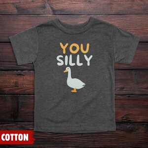 K&ouml;nnte beinhalten: Ein dunkelgrau meliertes Kurzarm-T-Shirt mit dem Text YOU SILLY in gelben und wei&szlig;en Buchstaben &uuml;ber einer Grafik einer wei&szlig;en Gans mit orangefarbenem Schnabel und F&uuml;&szlig;en. Das T-Shirt ist als 100% Baumwolle gekennzeichnet.