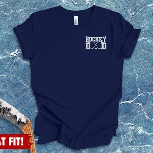 May include: A navy blue short-sleeve t-shirt featuring a white graphic on the upper left chest that reads HOCKEY DAD with two crossed hockey sticks positioned between the letters D and A. The shirt has a crew neckline and a standard fit.
