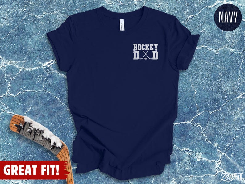 May include: A navy blue short-sleeve t-shirt featuring a white graphic on the upper left chest that reads HOCKEY DAD with two crossed hockey sticks positioned between the letters D and A. The shirt has a crew neckline and a standard fit.