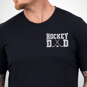 May include: A black short-sleeved t-shirt featuring a white graphic on the left chest. The graphic displays the text HOCKEY DAD in a block font, with two crossed hockey sticks and a puck replacing the letter A in the word DAD.