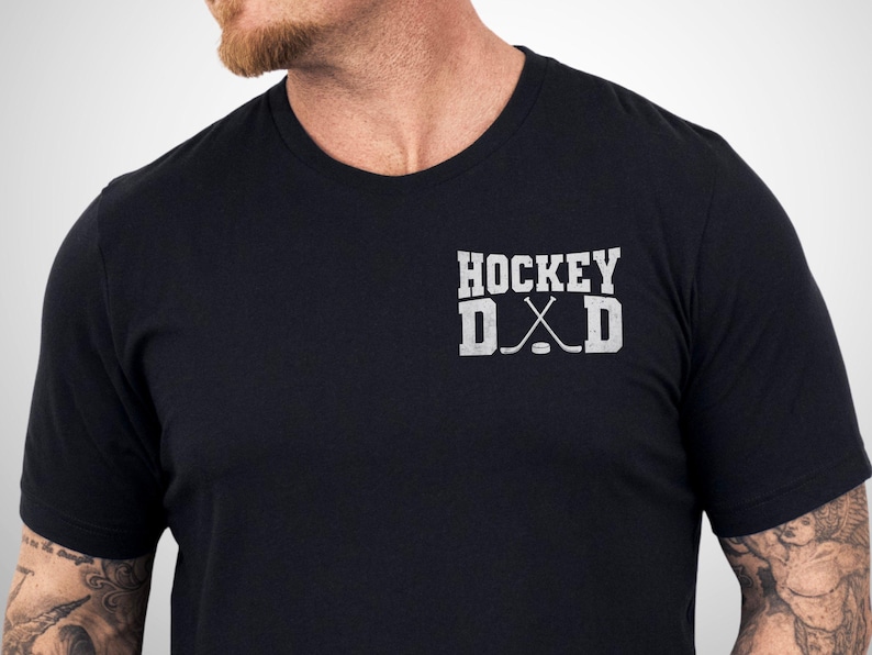 May include: A black short-sleeved t-shirt featuring a white graphic on the left chest. The graphic displays the text HOCKEY DAD in a block font, with two crossed hockey sticks and a puck replacing the letter A in the word DAD.