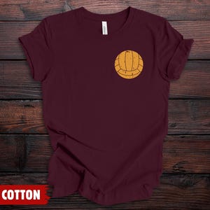 Puede incluir: Una camiseta de manga corta color granate con un peque&ntilde;o gr&aacute;fico de voleibol amarillo desgastado impreso en la parte superior izquierda del pecho. La prenda tiene cuello redondo y un corte est&aacute;ndar, mostrando un dise&ntilde;o sencillo de color s&oacute;lido.