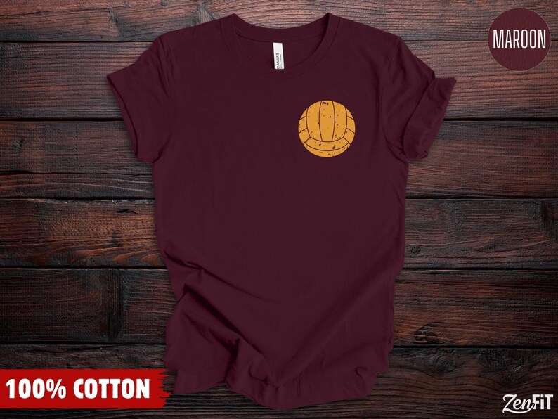 Puede incluir: Una camiseta de manga corta color granate con un peque&ntilde;o gr&aacute;fico de voleibol amarillo desgastado impreso en la parte superior izquierda del pecho. La prenda tiene cuello redondo y un corte est&aacute;ndar, mostrando un dise&ntilde;o sencillo de color s&oacute;lido.