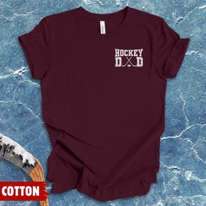 May include: A maroon short-sleeved t-shirt featuring the white text HOCKEY DAD with a graphic of two crossed hockey sticks and a puck. The shirt is made of 100% cotton and displays a simple crew neckline and a standard fit for casual wear.