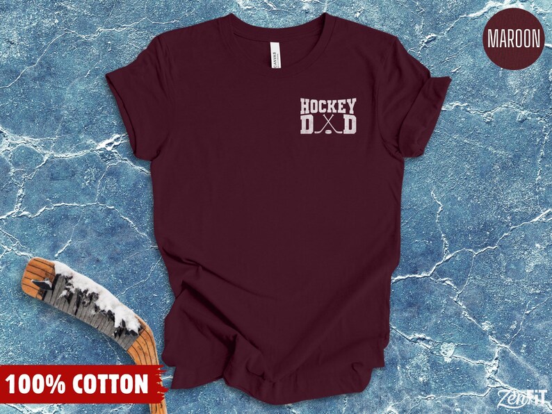 May include: A maroon short-sleeved t-shirt featuring the white text HOCKEY DAD with a graphic of two crossed hockey sticks and a puck. The shirt is made of 100% cotton and displays a simple crew neckline and a standard fit for casual wear.