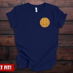 Puede incluir: Una camiseta de manga corta en color azul marino con un peque&ntilde;o gr&aacute;fico de un bal&oacute;n de voleibol amarillo desgastado situado en el pecho izquierdo. La prenda tiene cuello redondo y un corte est&aacute;ndar con un dise&ntilde;o sencillo de bal&oacute;n.