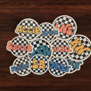 May include: A set of ten round stickers with a checkered black and white background. Each sticker features a different name in a colorful, bubbly font. The names include Allie, Lauren, LNS, Sarah, Ash, Achel, Kin, Mattie, SB, and Mily.