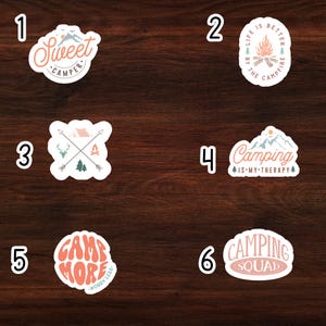 May include: Six different camping themed stickers on a wooden background. The stickers are all round and have different designs. The first sticker says "Sweet Camper". The second sticker says "Life is Better by the Campfire". The third sticker has a tent, two trees, and two deer. The fourth sticker says "Camping is My Therapy". The fifth sticker says "Camp More Worry Less". The sixth sticker says "Camping Squad".