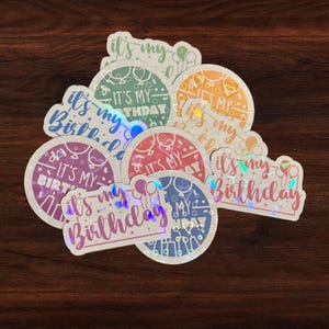 Puede incluir: Un conjunto de ocho pegatinas iridiscentes con el texto "It's my birthday" en varios colores y fuentes. Las pegatinas son redondas y tienen un acabado hologr&aacute;fico.