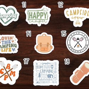 May include: Set of 18 camping themed stickers with various designs, including a heart with the word "Camping", a backpack, a campfire, and a marshmallow. The stickers are on a wood background.