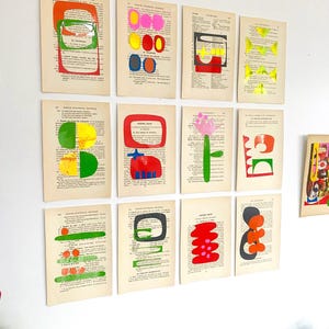 Op de afbeelding: Een verzameling van twaalf rechthoekige vintage woordenboekpagina's gerangschikt in een raster, elk met unieke abstracte vormen geschilderd in levendige kleuren zoals rood, groen, geel, roze en blauw, aangebracht over de gedrukte Franse tekst.