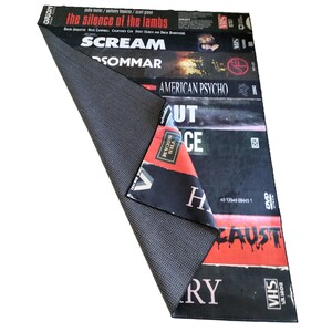 Może przedstawiać: Prostokątny czarny dywan z wzorem przypominającym kasety VHS. Widoczne są tytuły film&oacute;w, takie jak "The Silence of the Lambs" i "Scream". Dywan ma antypoślizgowy sp&oacute;d i zagięty r&oacute;g.
