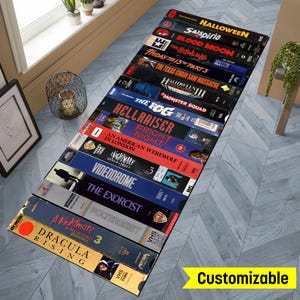 May include: A long, narrow rug featuring a design of VHS tapes with horror movie titles, including "Halloween", "Suspiria", "The Fog", "Hellraiser", "An American Werewolf in London", "Nightmare on Elm Street", "Videodrome", "The Exorcist", "Poltergeist", "A Nightmare on Elm Street 3", and "Dracula Rising". The rug is customizable.