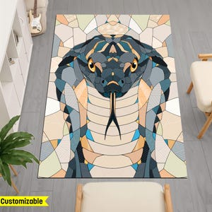 Op de afbeelding: Een rechthoekig tapijt met een geometrisch ontwerp van een cobra slangenkop. De slang is afgebeeld in tinten blauw, grijs en bruin, met gele ogen en een gespleten tong. Het tapijt heeft een mozaïekachtig patroon. Het woord "Aanpasbaar" staat linksonder.