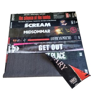 Może przedstawiać: Prostokątny dywan z motywem ułożonych kaset VHS. Widoczne tytuły to "The Silence of the Lambs", "Scream" i "American Psycho". Dywan ma czarne i szare tło i postarzany wygląd.