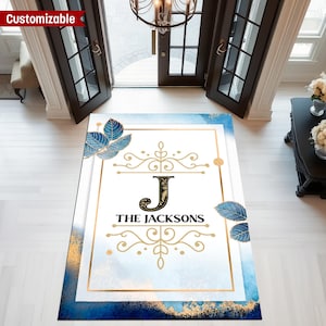 May include: Customizable rectangular doormat with a blue and gold watercolor design. The mat features a gold border, the letter "J", and the name "THE JACKSONS" in black. Blue leaf accents are placed on the corners.