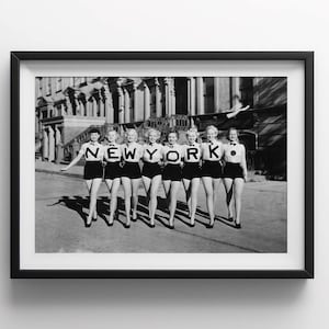 May include: Black and white photo of seven women holding letters that spell out "NEW YORK" in front of a row of buildings.