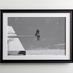 May include: Black and white framed print featuring a person sitting on a diving board. A seagull flies in the background over the water. The print is in a black frame with a white mat.