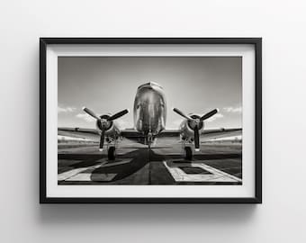 Peut inclure: Une photographie en noir et blanc d'un avion vintage sur une piste. L'avion fait face à l'appareil photo avec ses hélices visibles. L'avion est sur une piste avec une ombre projetée au sol.