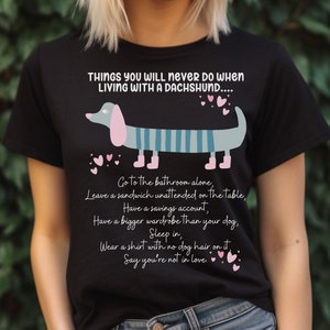 May include: Black t-shirt with a graphic of a dachshund and the text "THINGS YOU WILL NEVER DO WHEN LIVING WITH A DACHSHUND..."  The text lists humorous things you will never do when living with a dachshund, such as "Go to the bathroom alone" and "Sleep in".